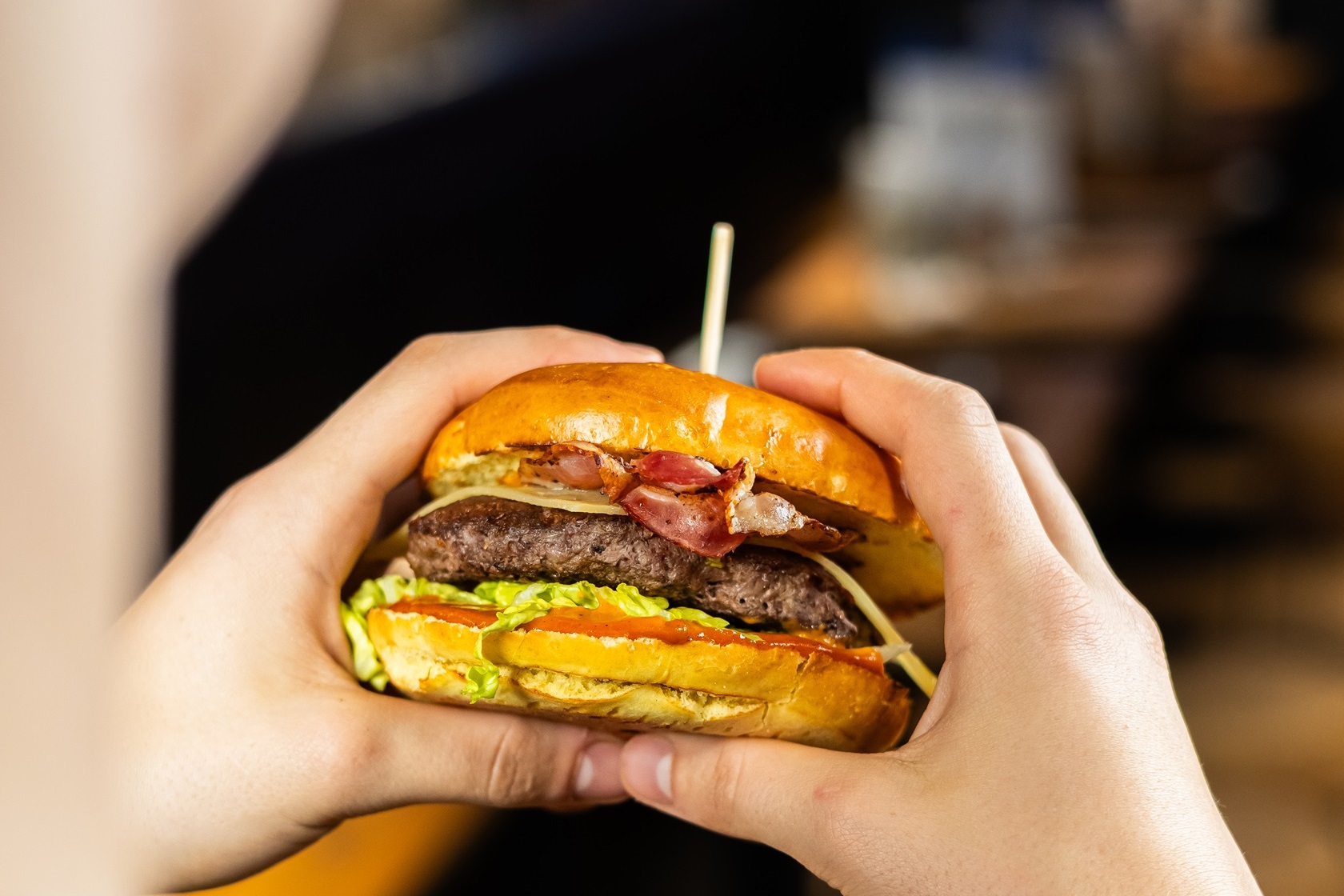 Eine Person hält einen Burger mit Bacon, Salat und einer Fleischscheibe in den Händen.